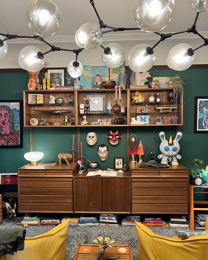 One wall of living room with large wood credenza with matching shelves above filled with sculptures, masks, other art objects. Dark green wall behind. We can see the backs of 2 yellow chairs and a ceiling light fixture that looks like a black tree branch with lights where fruits would be