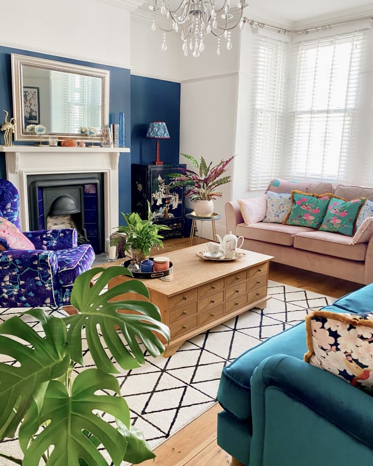 Living room with teal velvet sofa, pale pink velvet loveseat with aqua floral pillows, white walls, black and white geometric line rug, floral asian motif pillows, lamp, wood floor, pink accents, blue accent wall behind fireplace, crystal chandelier, velvet armchair in floral blue violet, large wood coffee table with drawers