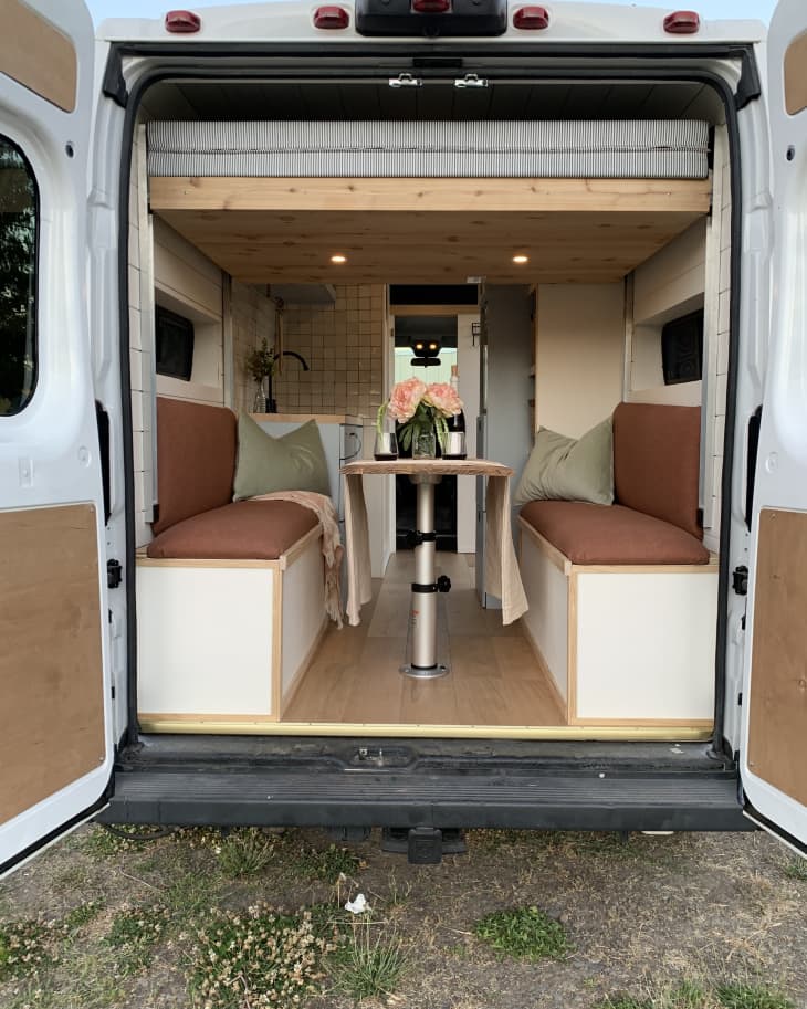 trunk view, bench seating with maroon bench cushions, table with adjustable pole in middle of benches, loft shelf for bed, sage pillows, view to front windshield, sink, black faucets, cabinets