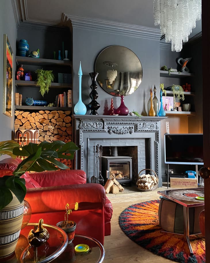 Living room with deep gray walls and fireplace, vintage white hanging light fixture, round sunburst shag rug, red leather loveseat, large stack of firewood and built in shelves with colorful glass vases and art objects