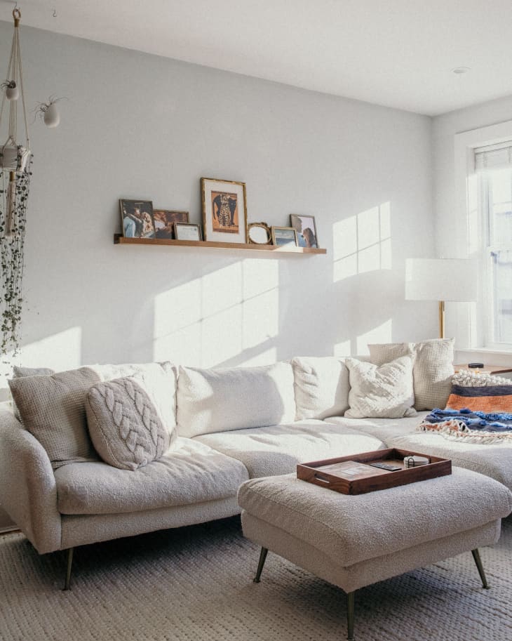 Cozy living room with a cream sectional sofa, ottoman, wall shelf with framed photos, and a floor lamp by the window.