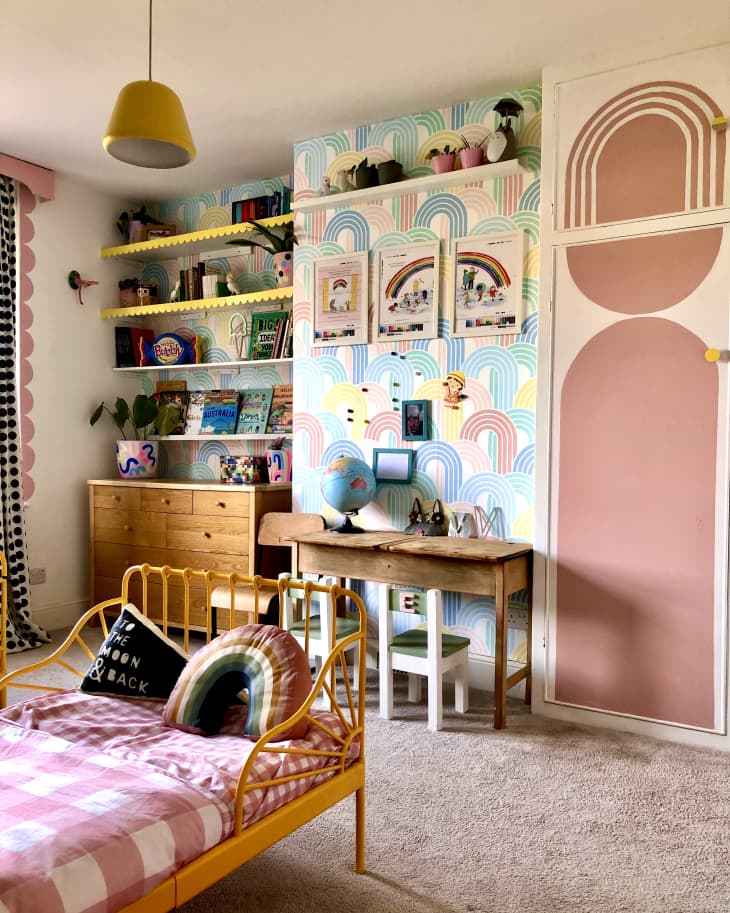 beige carpet, wooden dresser, scalloped yellow trim on book shelf, floating book shelves, globe, small wooden table, kids books, kids toys, scallop pink trim down window side, rainbow pattern wallpaper, plants, yellow metal bed frame with footer, pink and white checkered sheets, rainbow throw pillow, floor to ceiling closet doors, built in cabinets, dusty rose arched mural on closet doors