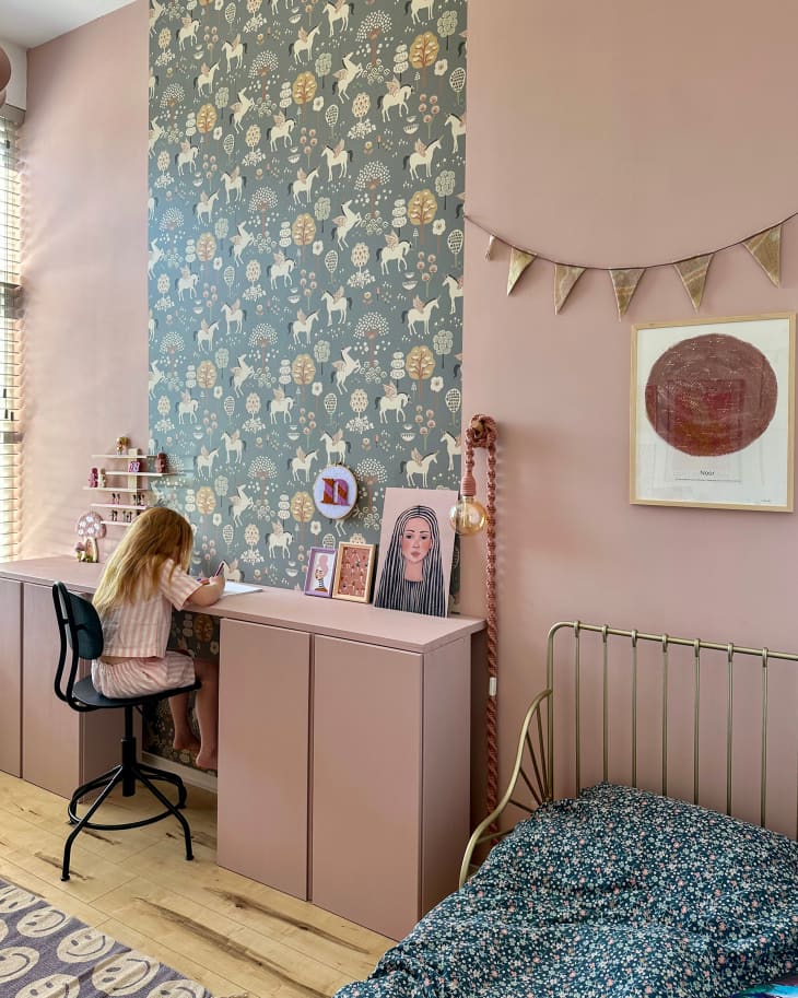 Small child seated at pink desk in pastel pink painted room with a floral and horse wallpaper strip going down wall in front of desk. Brass bed with floral bedding in corner.