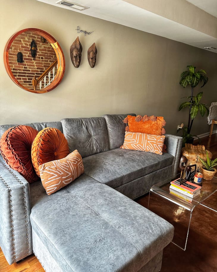 living room with gray sectional with orange throw pillows, round mirror on neutral colored wall, 2 masks next to it. Lucite clear coffee table, wood floors, plants