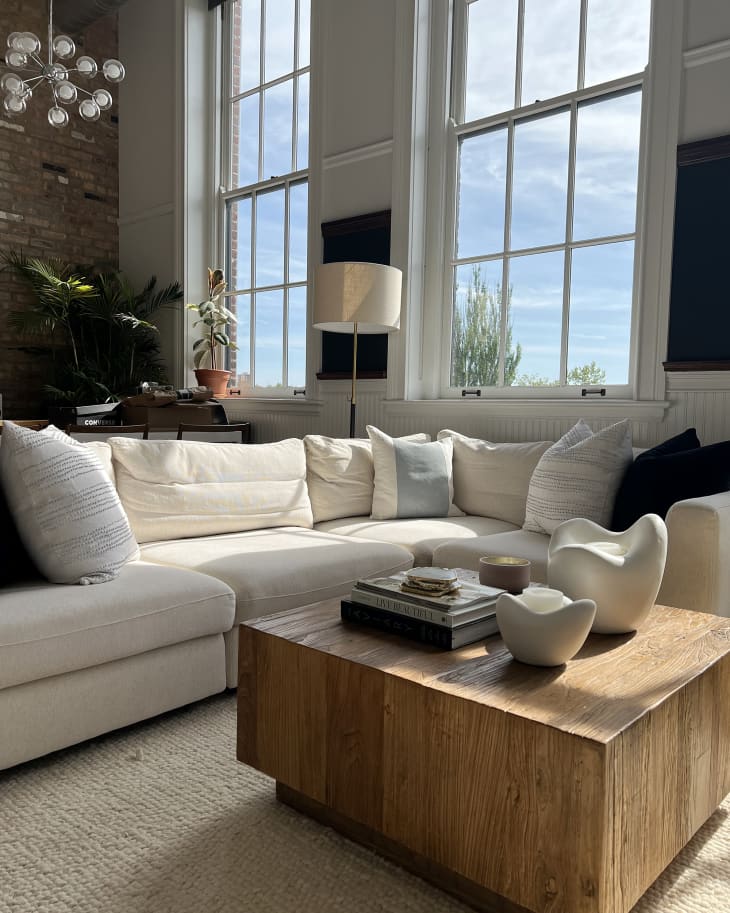 Living room with large white sectional sofa with lots of pillows, wood cube coffee table with white art objects, stack of books, white walls and one exposed brick wall, large, tall windows, natural color area rug