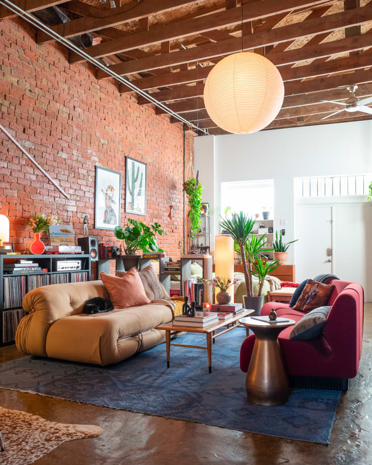 living room with high, exposed beam wood ceiling, white walls, lots of art (mostly paintings, some framed, some not) and gallery walls, large round hanging paper lantern, red and gold sofas facing each other on blue area rug, one exposed brick wall, work area in one corner