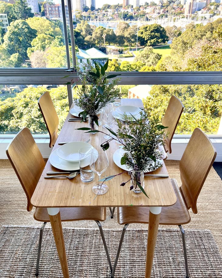 light wood rounded edge rectangular dining table with matching modern chairs on layered natural area rugs. Giant full-wall window behind. Table is set with white place settings, glassware. Vases of flowers
