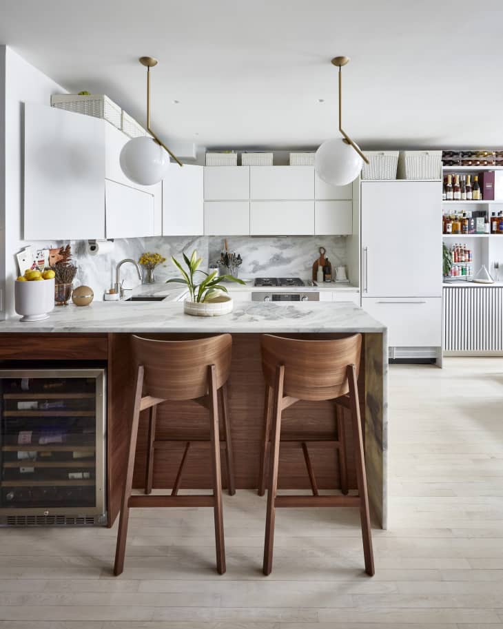 Kitchen with marble bar/counter/backsplash, curve-backed wooden stools, white cabinets, fridge, white globe pendant lights