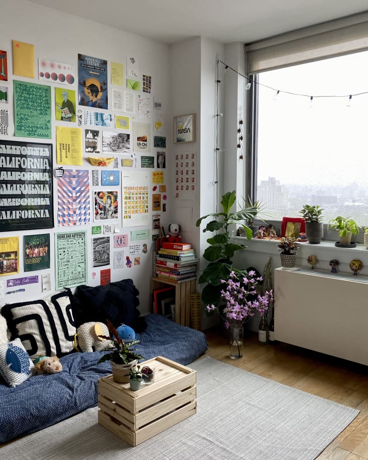 view from entryway into living space in brooklyn apartment: cushioned seating on floor with navy blue cover, black white and navy throw pillows. unframed art gallery wall on wall behind. crate as coffee table and crate with stack of books. Plants on floor and radiator, large window, white walls, pale neutral area rug