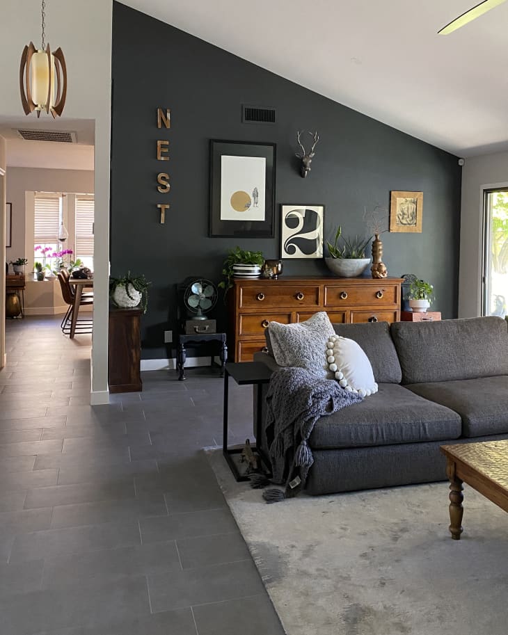 Living room with dark gray sofa, light gray rug, gray tiled floor, one dark gray accent wall, wood dresser with art above, angled ceiling, view into dining room