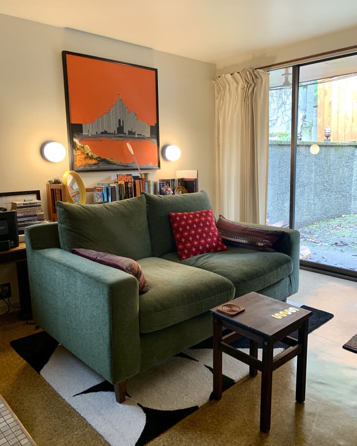 Living room with green/blue loveseat with throw pillows, black and white area rug, small table, large windows, art on wall behind