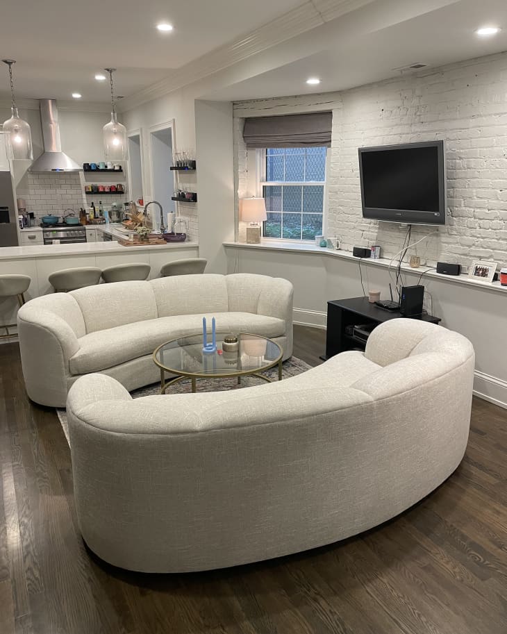 Living room with exposed white brick wall, white shelf along entire wall, under TV, 2 white curved sofas, wood floor, round glass and gold coffee table, kitchen in background