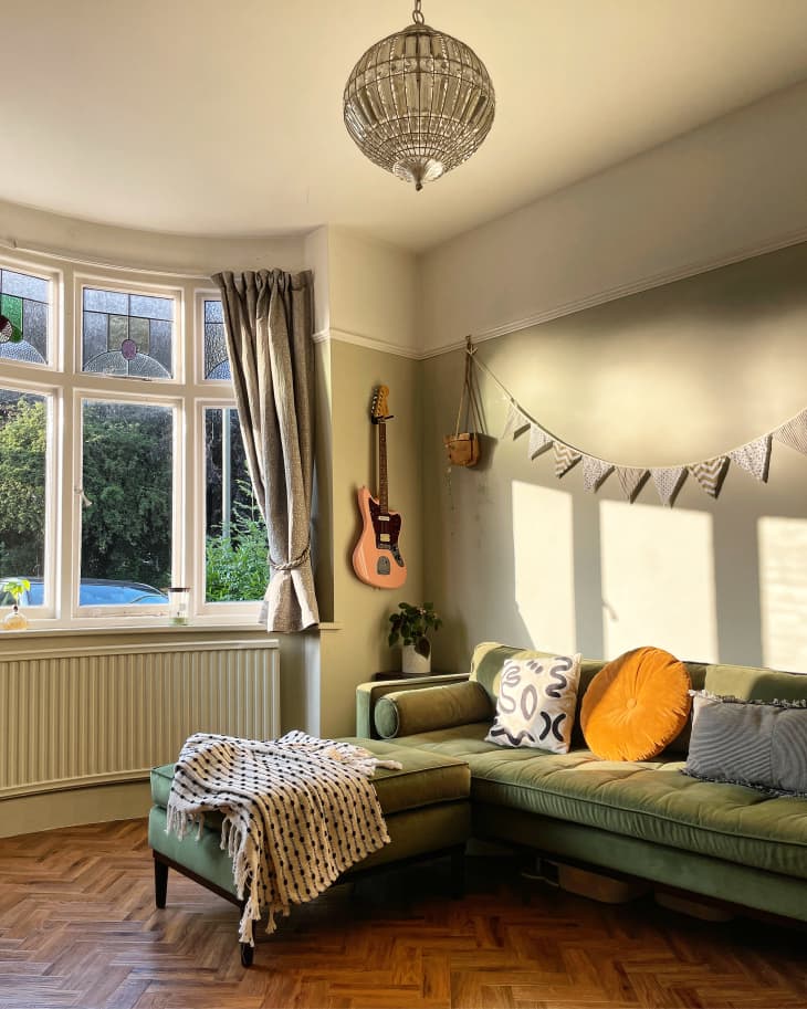 Green sectional sofa with golden hour light and guitar on the wall, herringbone wood floor, stained glass upper bay windows