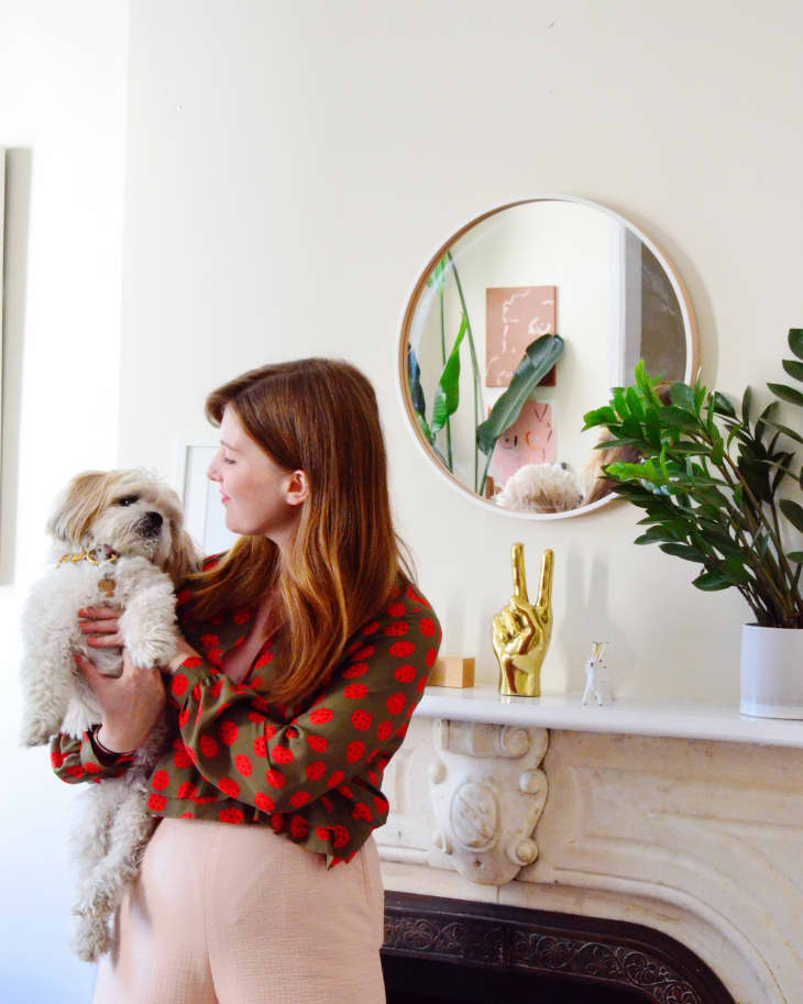 Woman holding a dog in front of a fireplace with plants, a round mirror, and a gold peace sign sculpture.