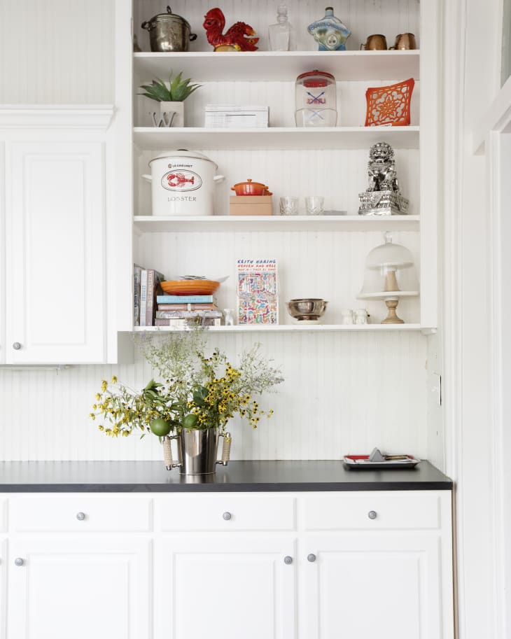 White kitchen cabinets with black countertop, open shelves displaying colorful decor, and a vase of yellow flowers.