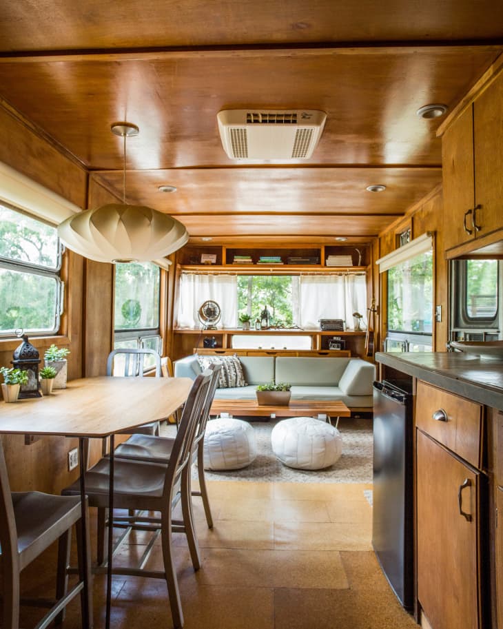 Retro camper interior with wooden paneling, a light green sofa, dining table, chairs, and poufs, surrounded by large windows.