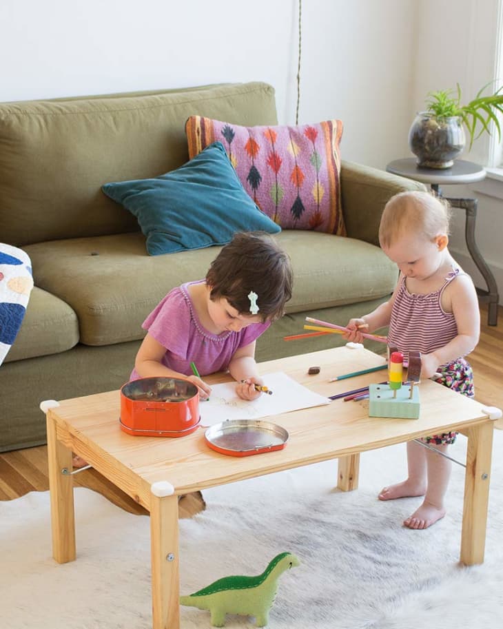 Two children drawing at a wooden table in a living room with a green sofa and colorful pillows.