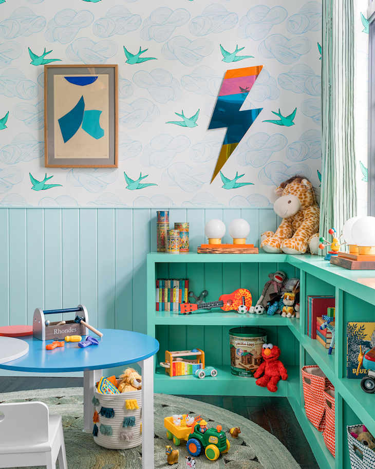 Light blue tongue and groove half wall paneling with cloud illustrated wallpaper with teal sparrows, light teal low built bookshelves in L shape, kids play table