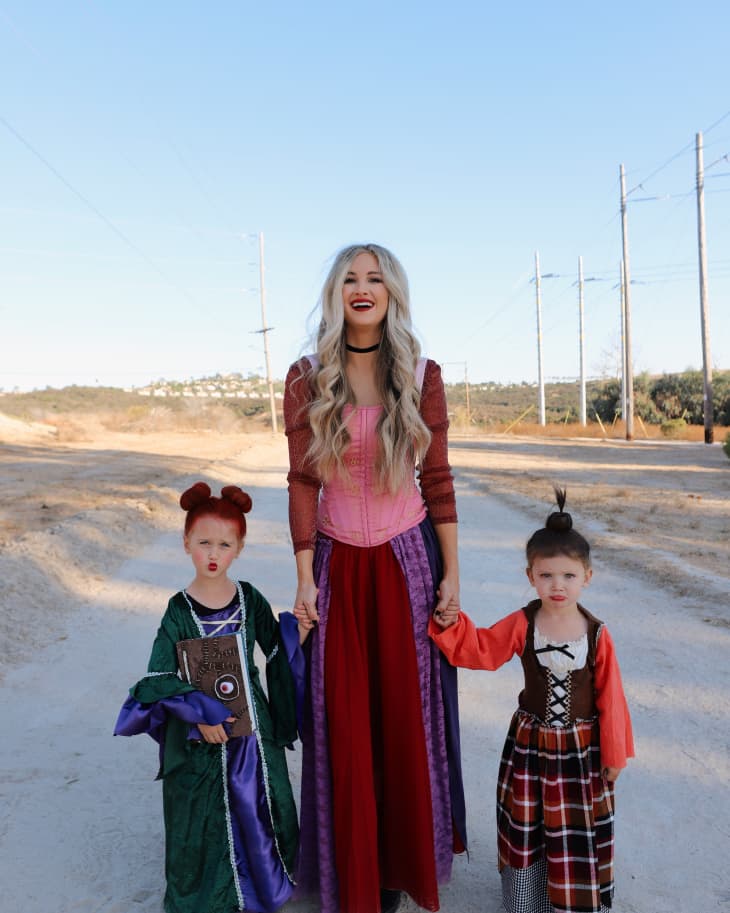 Woman and two children in colorful Halloween costumes standing outdoors on a dirt path.