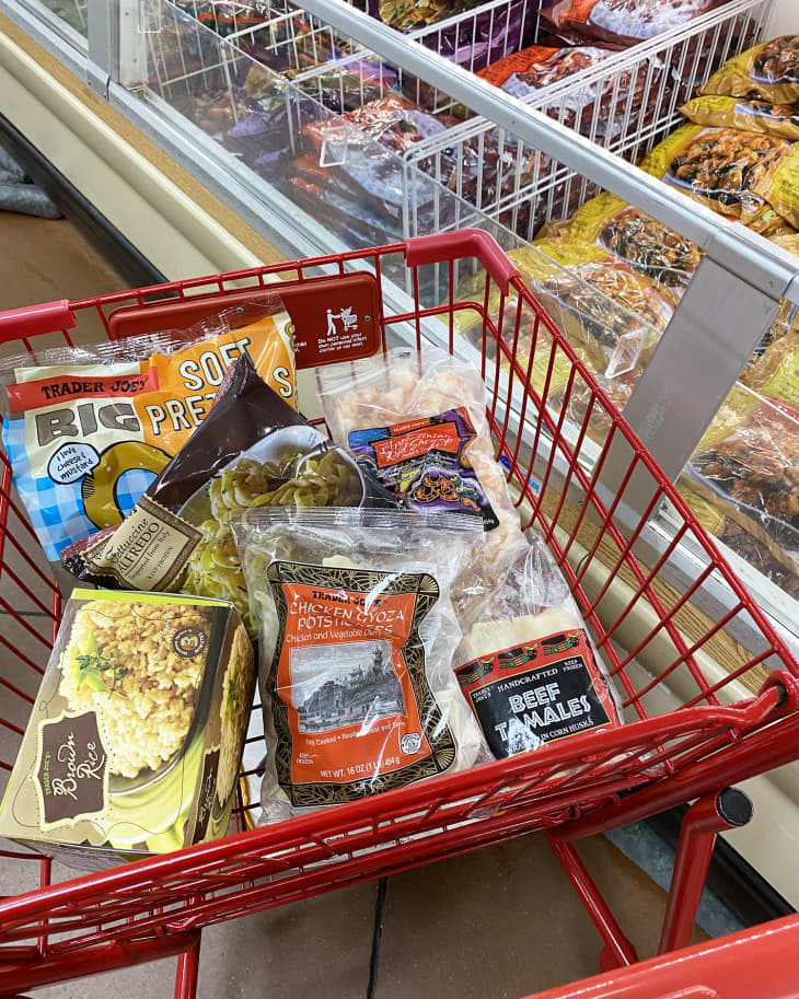 Shopping cart with frozen foods including chicken gyoza, beef tamales, soft pretzels, and boxed rice near freezer aisle.