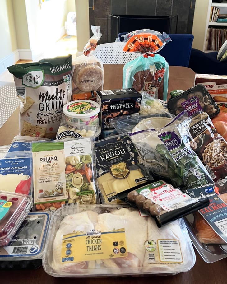 Assorted groceries on a table, including bread, cheese, chicken thighs, salmon, ravioli, and fresh produce.