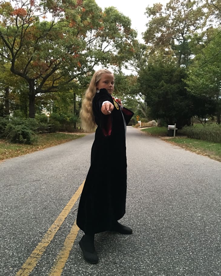 Child in a wizard costume with a wand, standing on a tree-lined road.