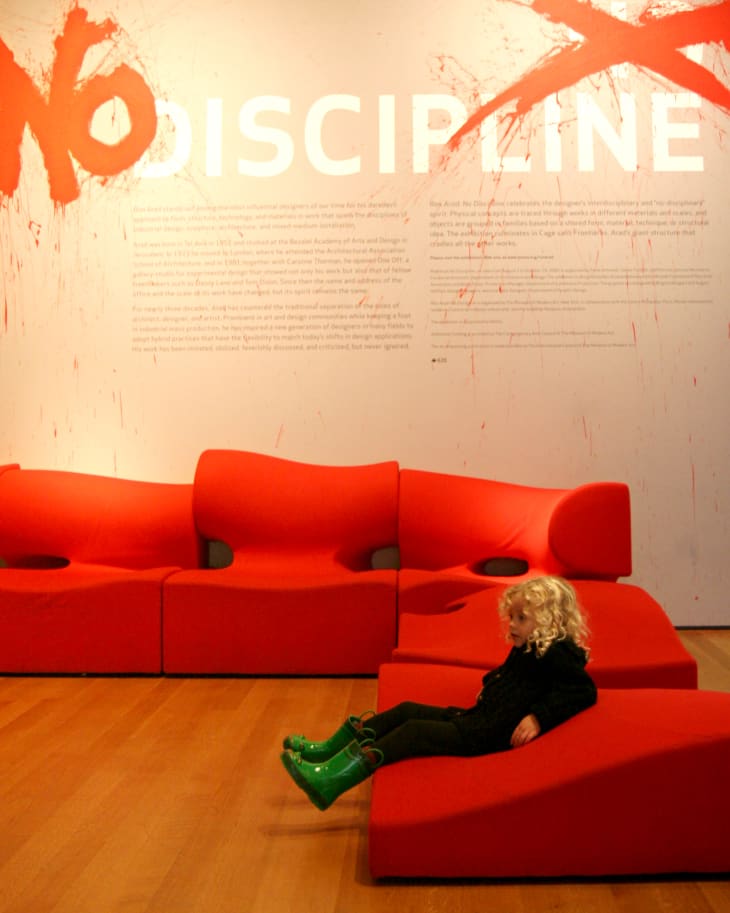 Child in green boots sitting on a modern red sofa with "No Discipline" text on the wall behind.
