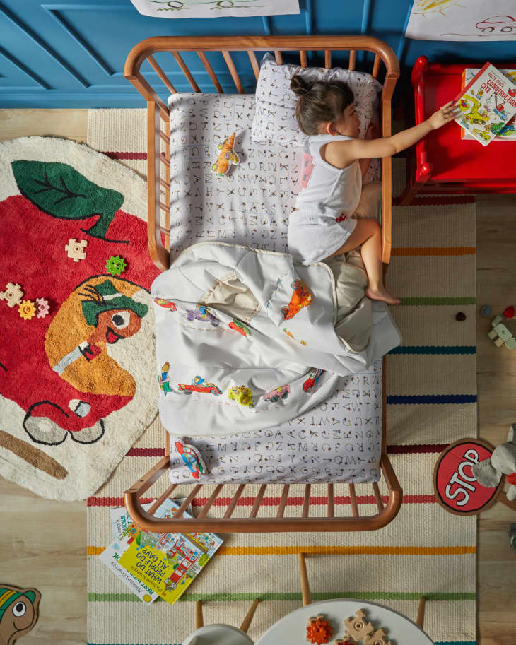 Wood bed frame with colorful striped rug on cream background, apple car rug and car quilt on bed, red nightstand, and blue wainscoting on wall