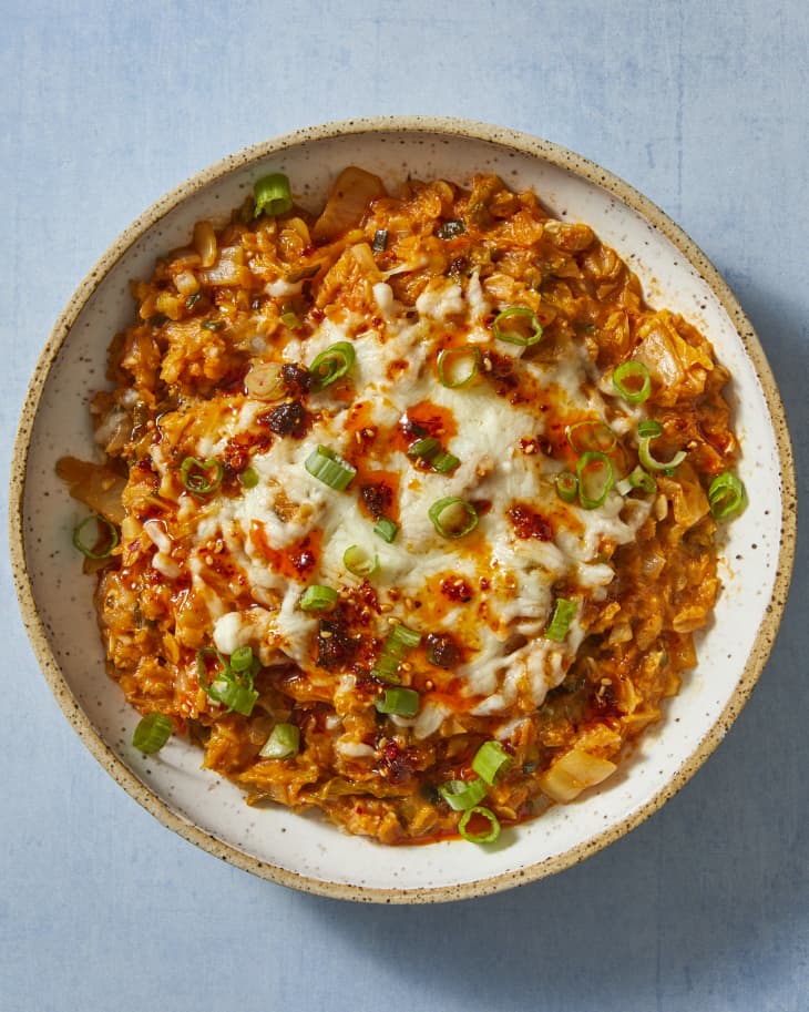 An overhead view of a white speckled stoneware bowl with savory kimchi oatmeal covered in melted cheese and scallions on a light blue surface.