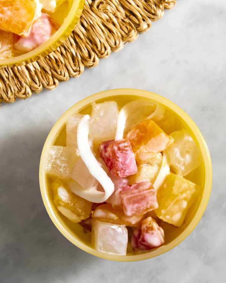 A yellow bowl of Filipino fruit salad with chunks of tropical fruit, coconut and condensed milk on a marble background with a rattan placemat peeking in