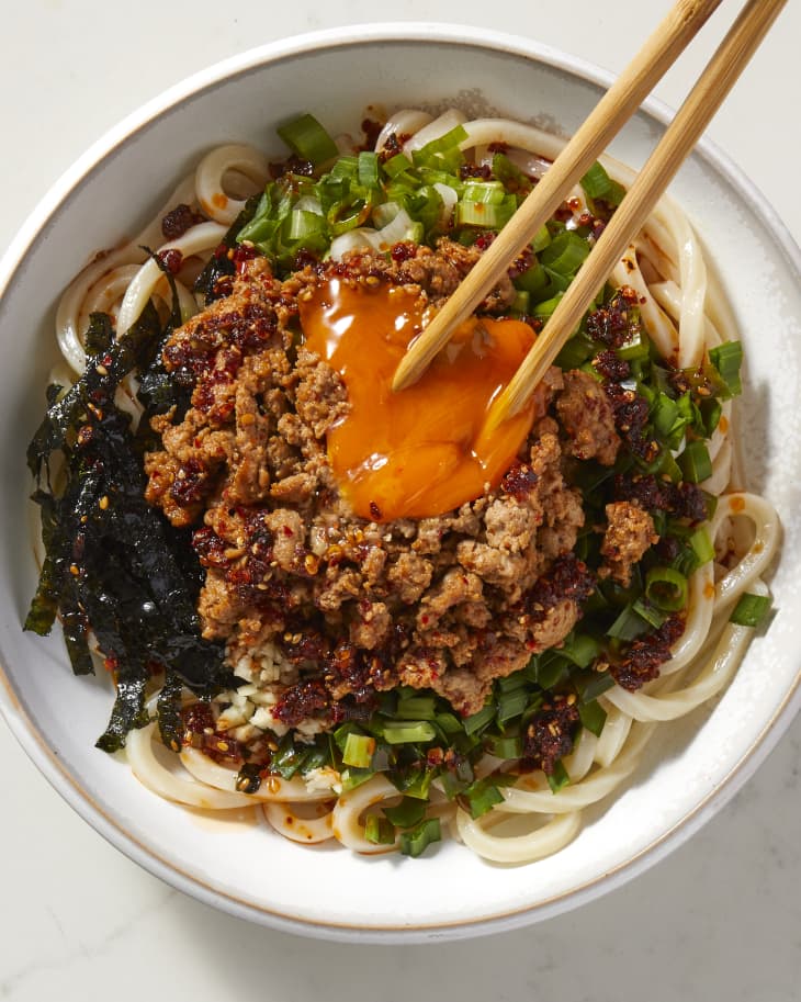 Overhead view of a bowl of chile crisp mazesoba with a bright orange egg yolk being broken with chopstick, toppings neatly placed and not mixed in, on a marble surface