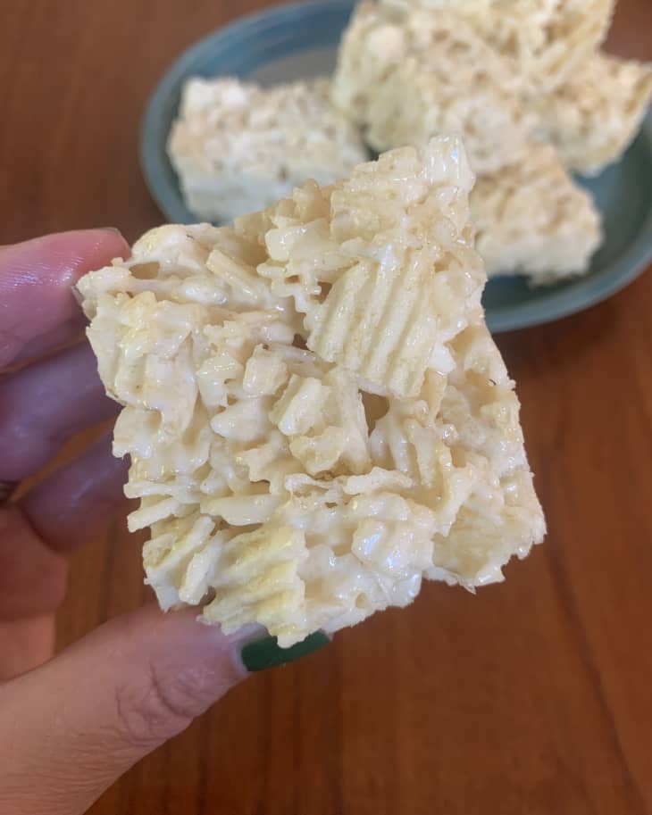 Close-up of a hand holding a square rice cereal treat with a plate of treats in the background.