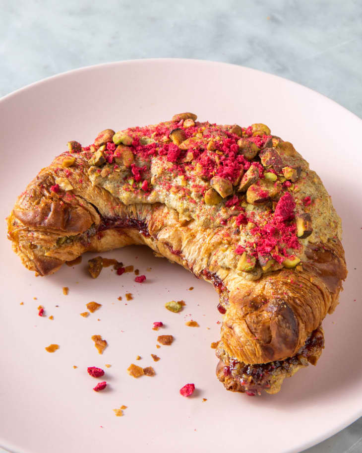 single pistachio raspberry croissant with crumbs of pistachio and raspberry next to it on a pink plate placed on a marble surface