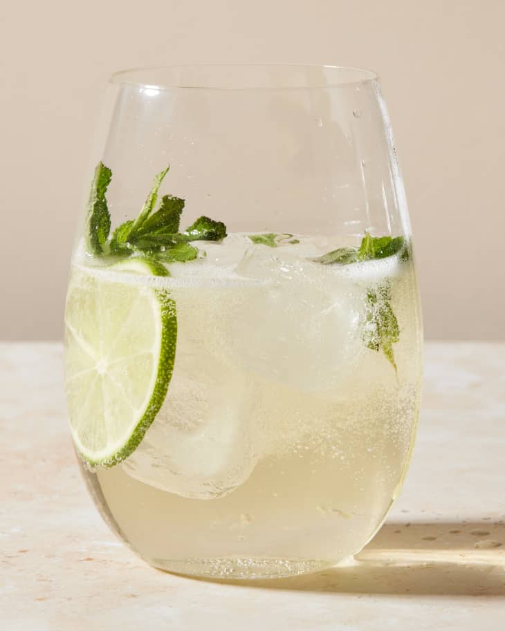 a semi wide shot of a glass of a hugo cocktail with lime and mint that's on rustic sandy colored surface