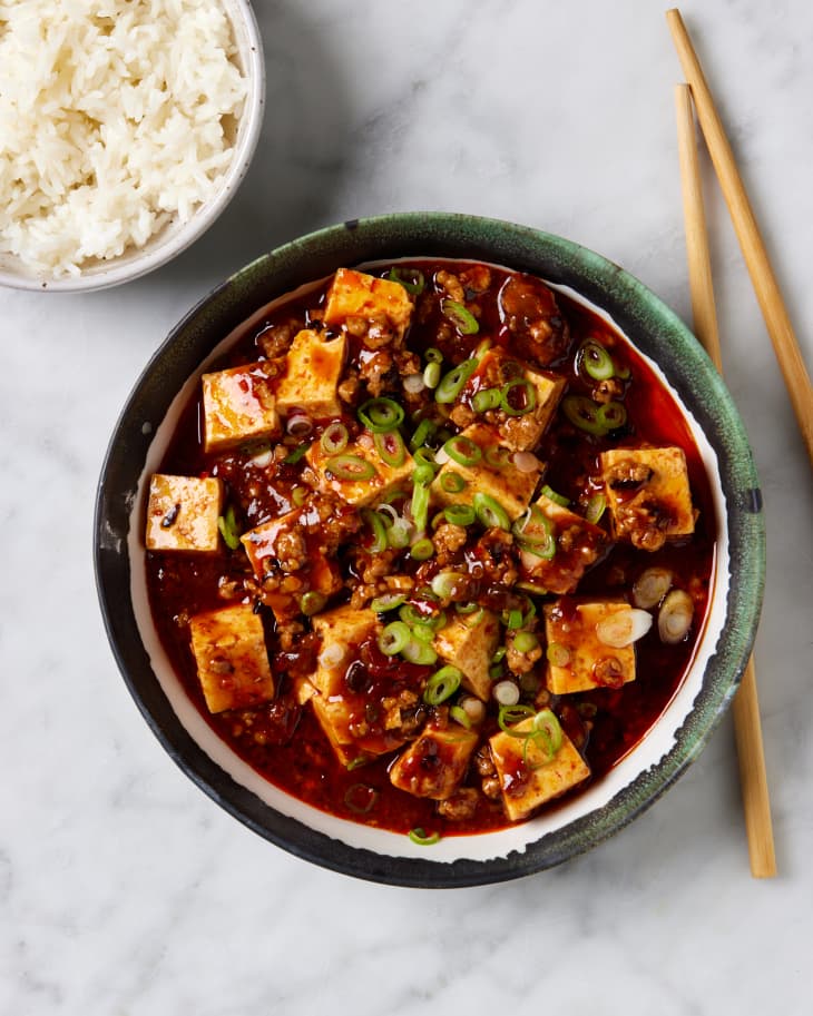 semi wide shot of mapo tofu in a green and white bowl with spring onions on top. chopsticks lay next it on the right hand side as well as a white bowl of white rice on the upper left hand side on a marble surface