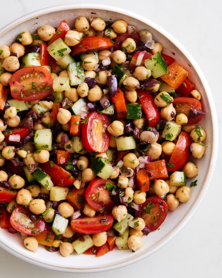 bowl of a mixed chickpea salad with chopped olives, red onions, red bell pepper,cucumber, and halved grape tomatoes on a marble surface