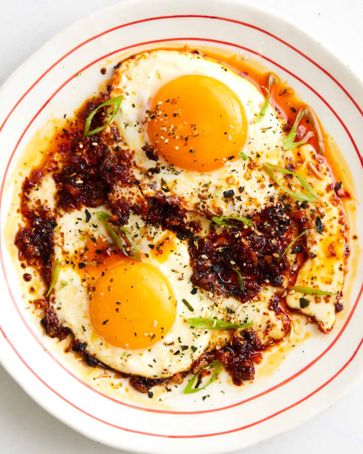 two sunny side up eggs with chili crisp on a white plate with red concentric circles, garnished with sesame seeds and scallions
