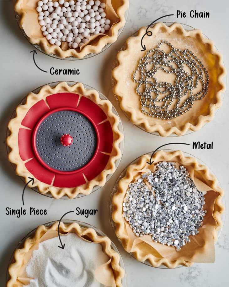 five different pie weights made of different materials shown in pie dishes and a pie crust on a marble surface