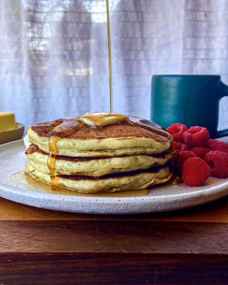 someone is drizzling syrup on. pancakes  on a plate next to fruit