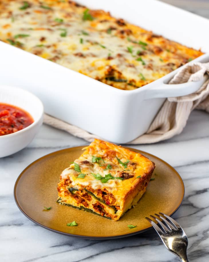 Photograph of Chile Relleno Casserole in baking dish and one slice on a plate with side dish of sauce on a marble countertop.