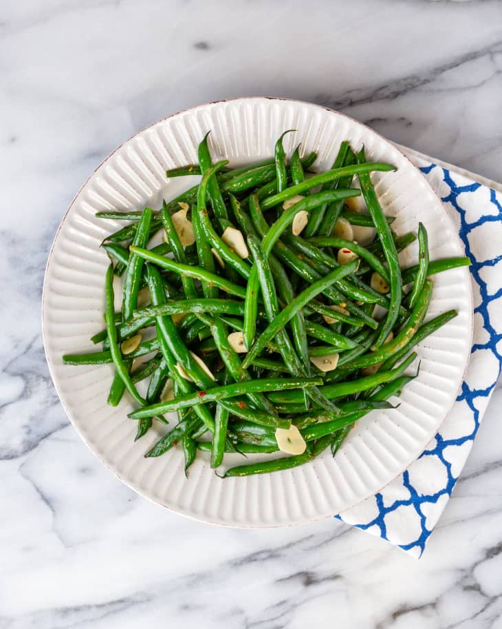 Photograph of garlic green beans on a white plate with kitchen towel on a marble countertop.