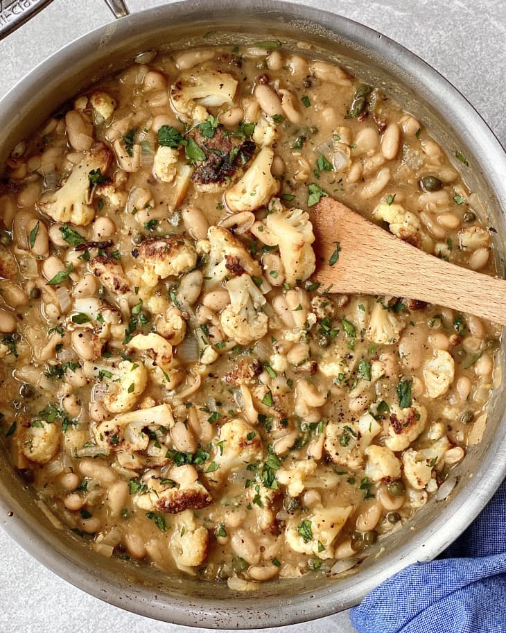 Braised cauliflower and white bean piccata in skillet with wooden spoon inside.