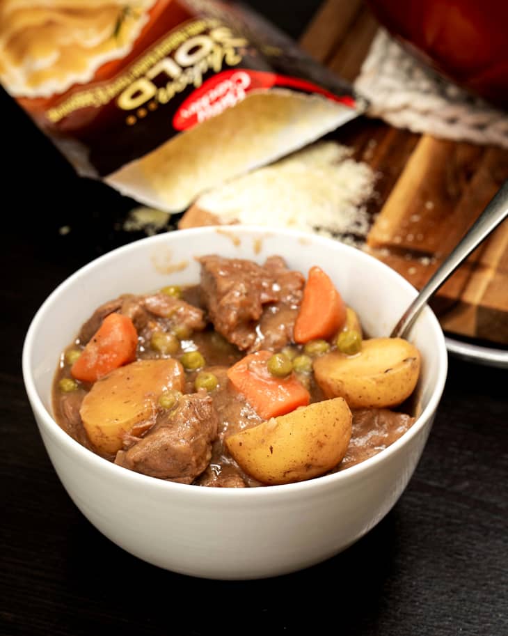 Beef, potato, carrot and pea stew that has been thickened by adding crushed potato chips in a white bowl with a spoon. The bag of chips is in the background, with crumbs spilling out