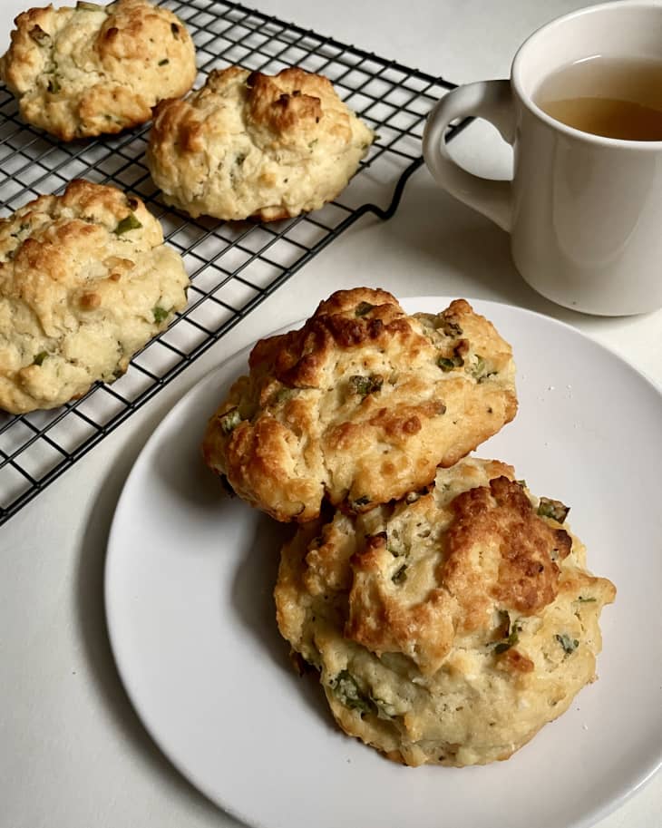 Photo of scallion cream cheese scones, some cooling on rack, one on a plate. cup of tea in upper right
