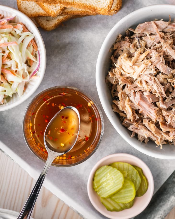 Carolina BBQ sauce in a bowl with pulled pork, white bread, cole slaw and pickles laid around.