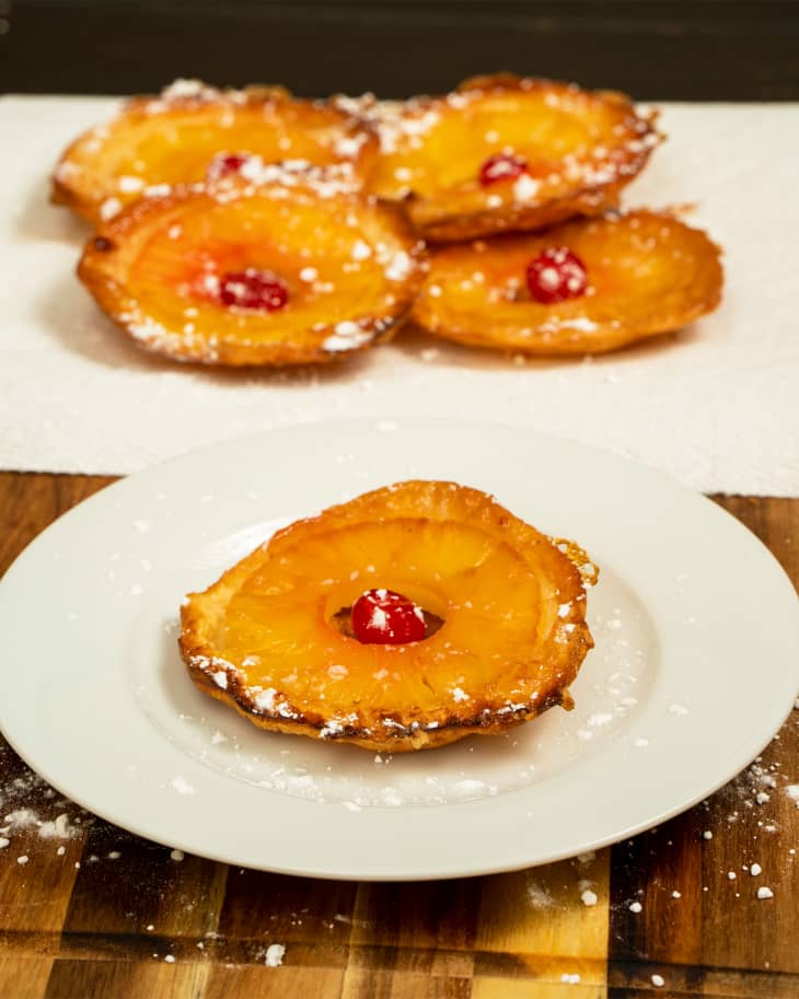 Powdered sugar dusted pineapple ring filled with cherry on a plate. Remaining cherry filled pineapples in background on paper towel.