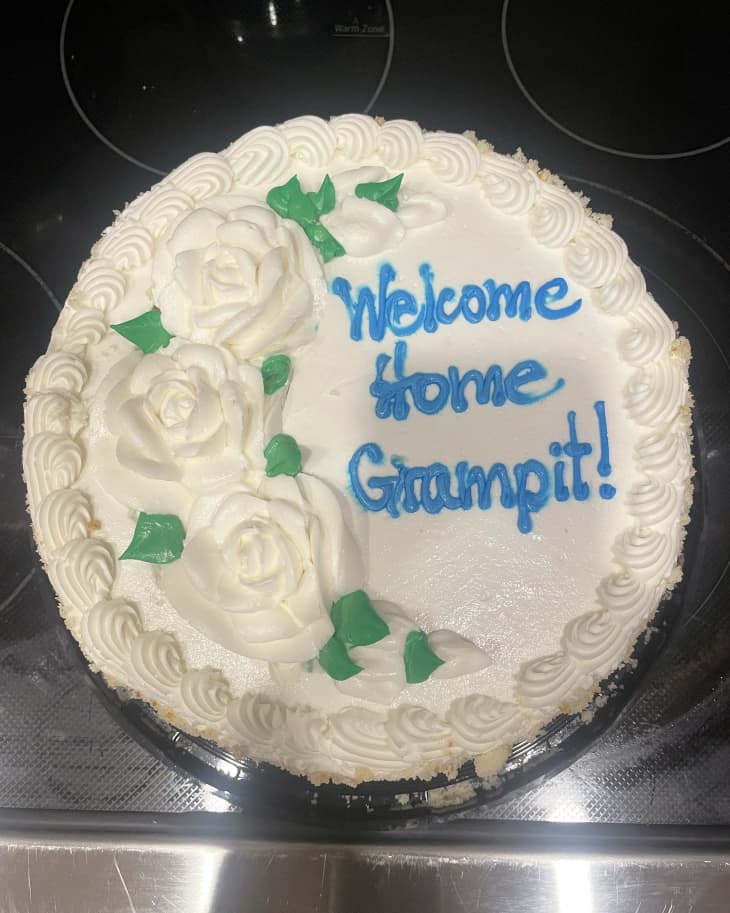 White round cake decorated by costco with white frosting flowers, green leaves, text says "Welcome Home Grampit"