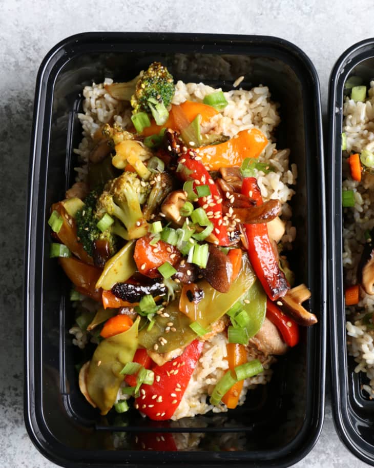 Stir-fried vegetables with rice in a black meal prep container, topped with sesame seeds and green onions.