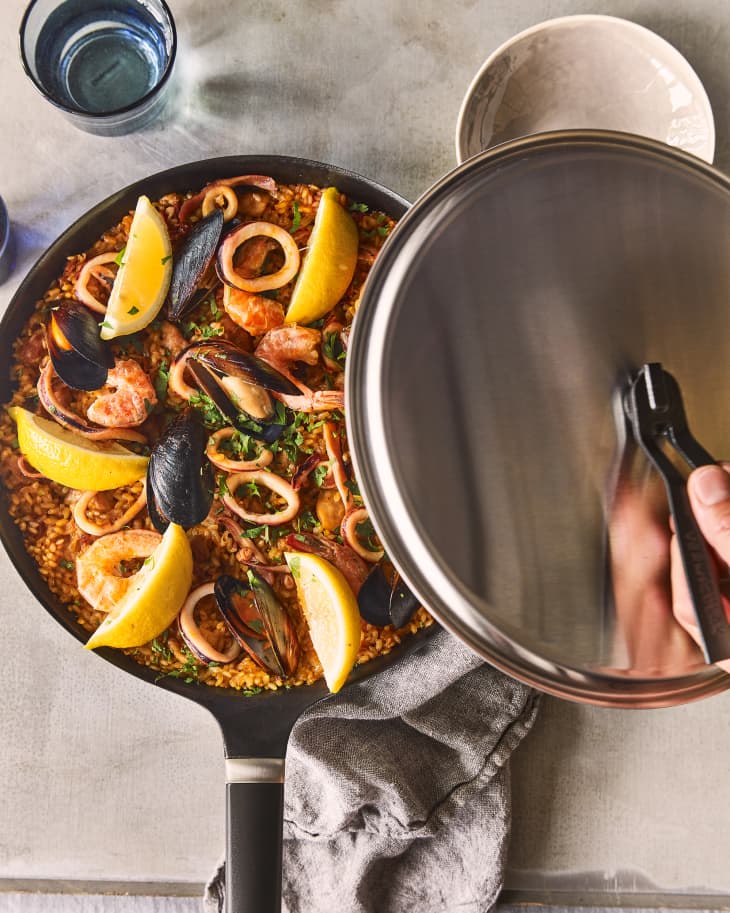 Vermicular’s Oven-Safe Skillet on countertop with seafood recipe and lemon wedges in it. A hand is lifting off the lid.