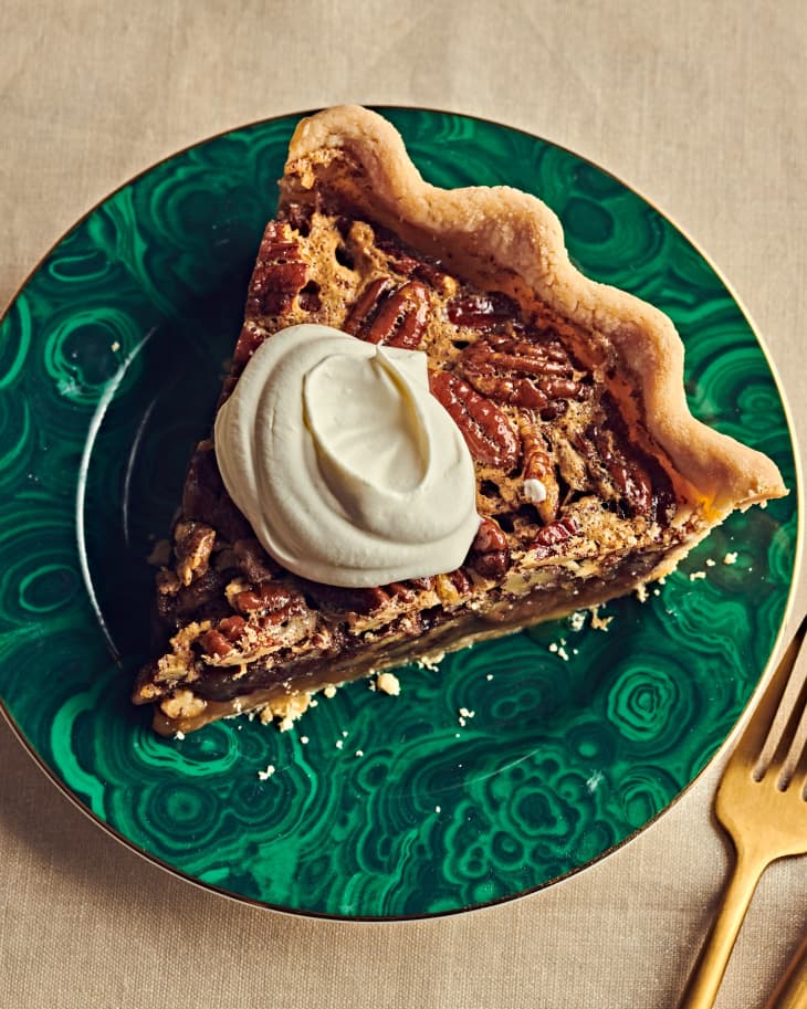 Slice of pecan pie on green plate with gold fork. Pie has a dollop of creme fraiche on top
